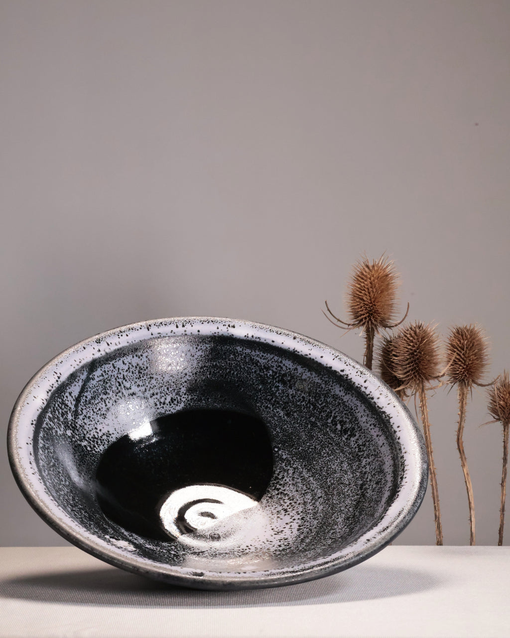 Black speckled ceramic bowl on a neutral background with dried plants.