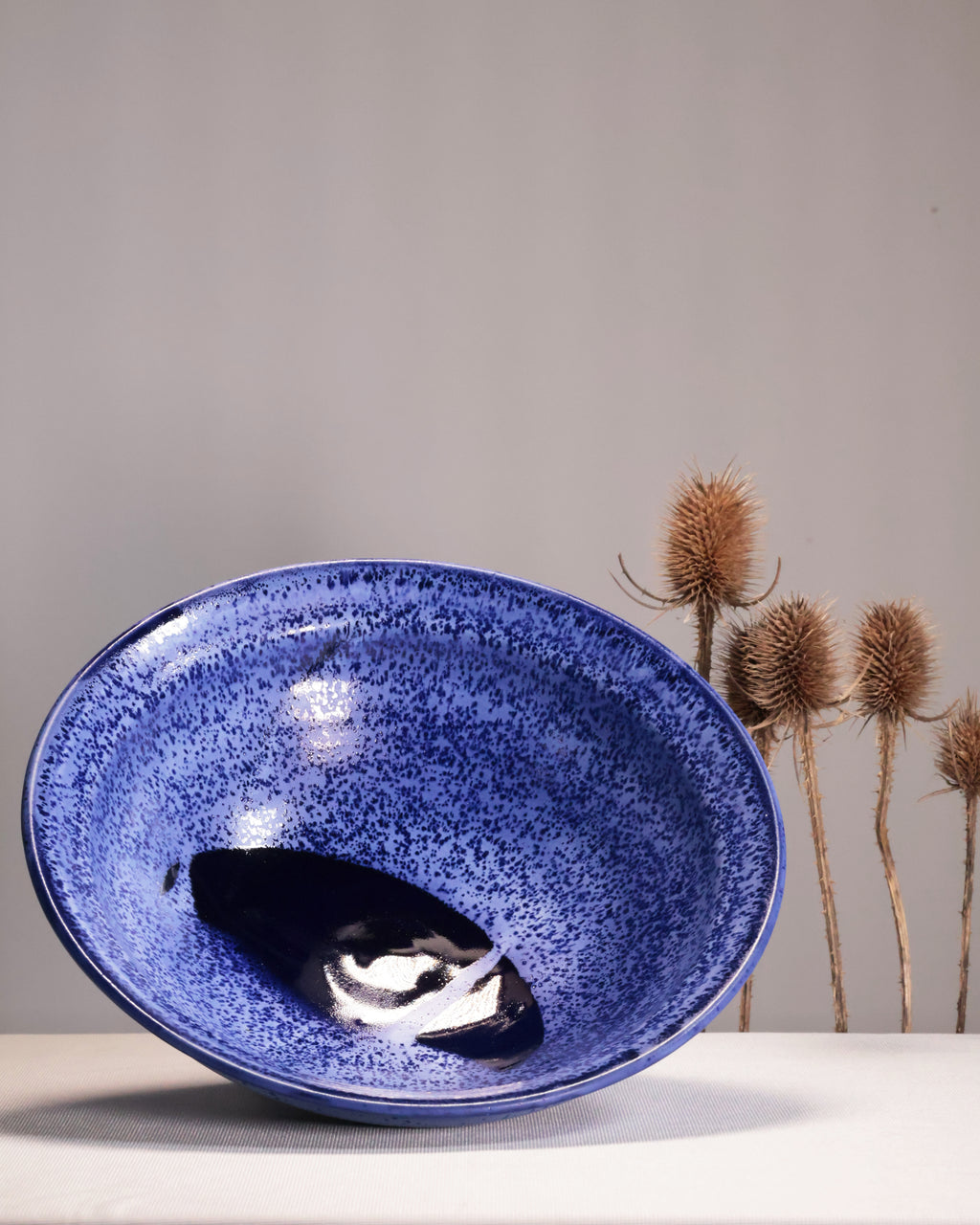 Blue ceramic bowl on a white surface with a gray background and dried plants.