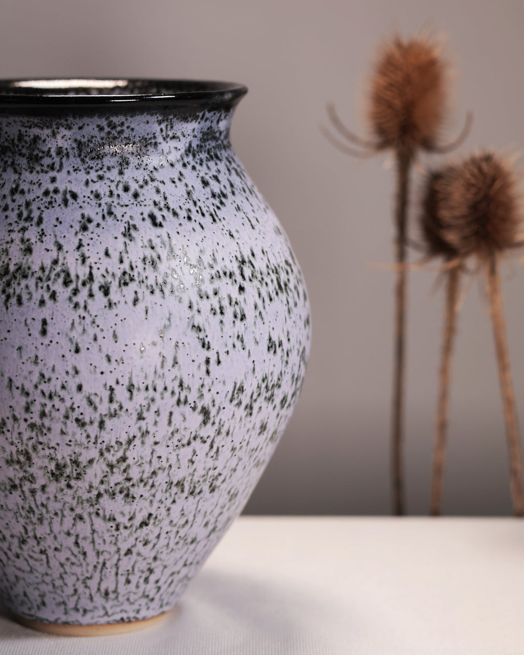 Speckled ceramic vase on a neutral background with dried plants.