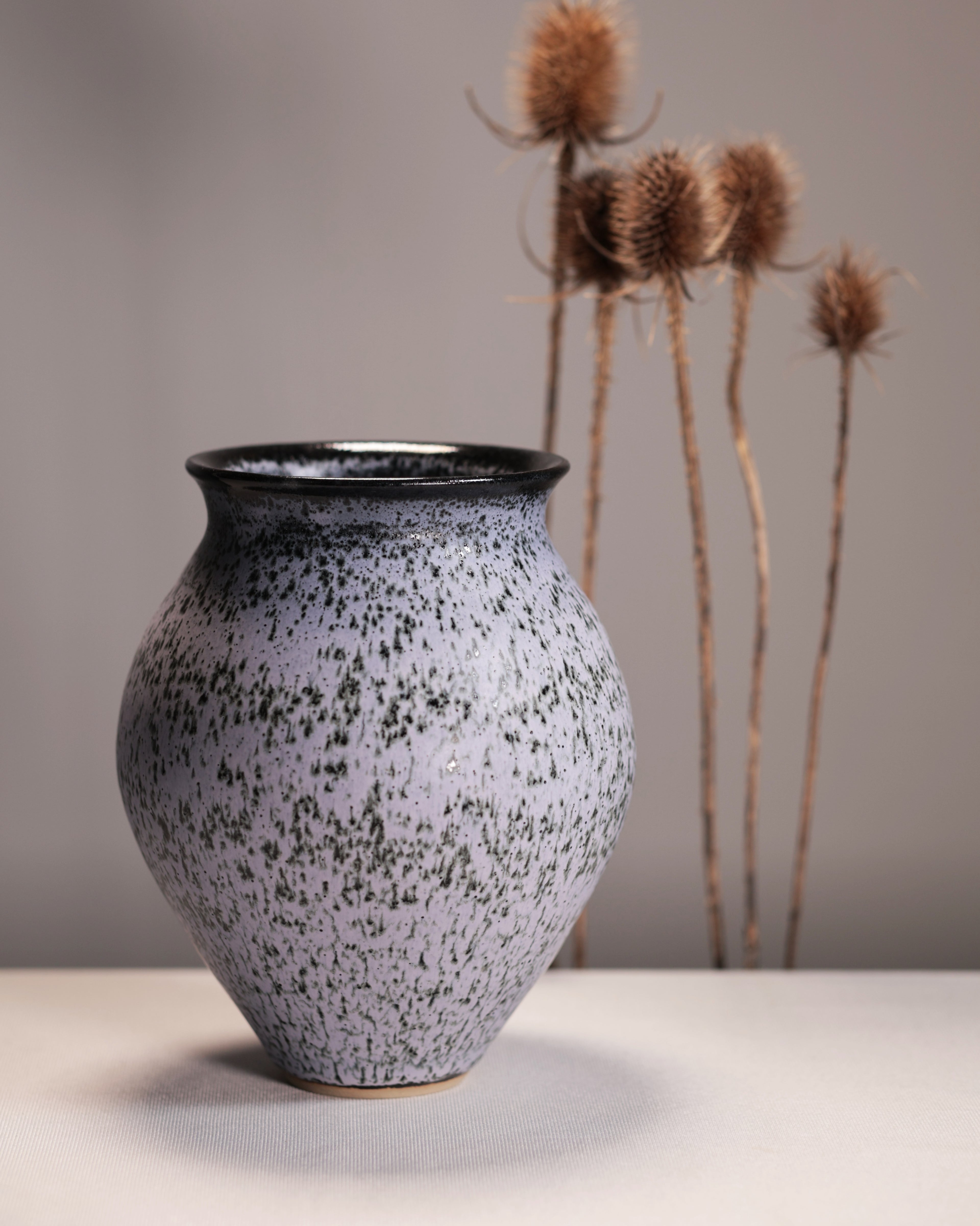 Speckled ceramic vase on a neutral background with dried plants.